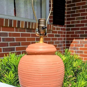 May include: A terracotta-coloured table lamp base with a textured, horizontally ribbed surface. The lamp features a rounded, bulbous body, a narrow neck, and a metallic brass-toned socket assembly with a harp attached to the top for a lampshade.