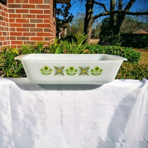 Puede incluir: Una fuente rectangular de vidrio blanco con un estampado floral en verde y amarillo. La fuente tiene asas en cada extremo. El diseño floral se repite en la parte delantera de la fuente. La fuente está sobre una superficie blanca.