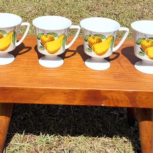 May include: Four white pedestal mugs with a yellow pear and green leaf design, arranged on a small, wooden, mid-century style table. The table has a rounded top and three legs. The mugs are on a grassy surface.