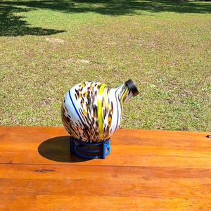 May include: A decorative glass bottle with a black spout and a colorful pattern of yellow, brown, and blue stripes. The bottle sits on a blue stand on a wooden surface.