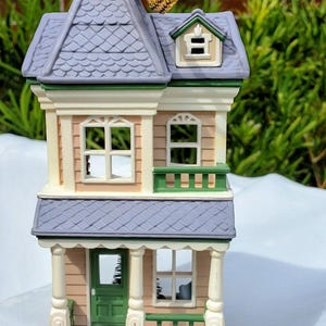 May include: A two-story house ornament with a lavender roof, tan siding, and a green front door. The structure features white trim, a small balcony, and a red front step. The year 1987 is printed vertically on the front porch pillar, and a gold cord is attached to the top for hanging.