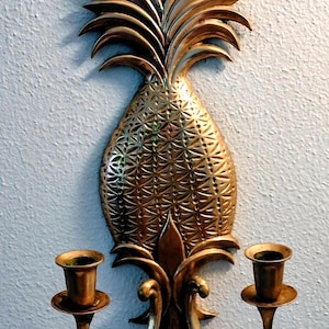 May include: A gold-colored wall sconce shaped like a pineapple, with two candle holders. The pineapple has a textured surface and detailed leaves. The sconce is mounted on a light blue wall.