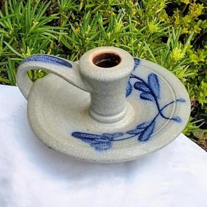 Puede incluir: Un candelero de gres hecho a mano con una base circular y un asa. El candelero es de color gris claro con diseños florales azules y un interior marrón. El asa es curva y el candelero está sobre una superficie blanca.