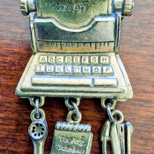 Può includere: Una spilla in metallo color oro a forma di macchina da scrivere con il testo APPOINTMENT 9:00AM e una tastiera con le lettere dalla A alla P. Tre ciondoli pendono dal basso: una cornetta del telefono, un blocco note con TODAY'S SCHEDULE e una cucitrice.