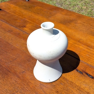 May include: A white ceramic vase with a rounded body and a flared base. The vase has a small, cylindrical opening at the top. The vase is sitting on a wooden surface.