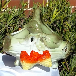 May include: A ceramic planter shaped like a stylized artichoke, in light green and white. It has an orange and yellow bow-tie design on the front. The planter is set against a backdrop of green foliage, with a hint of a brick wall.