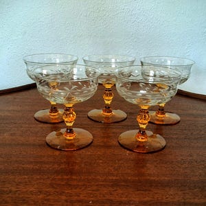 May include: Five vintage champagne coupe glasses with etched floral patterns. The glasses have amber-coloured stems and bases, and are arranged on a dark wooden surface. The stems have a decorative, beaded design.