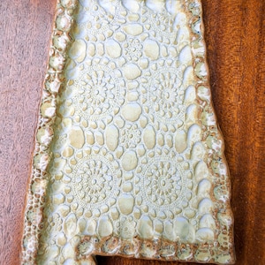 May include: A ceramic tray shaped like the state of Alabama with a light cream and pale green glaze. The surface features a textured, embossed pattern of circular lace-like motifs and organic pebble shapes, framed by a thick, uneven, bumpy border.
