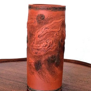 May include: A cylindrical, red-orange vase with a detailed dragon relief carving. The vase has a dark, mottled texture and a decorative band at the top. The base has a green and black patterned border. The vase is displayed on a dark wooden surface.