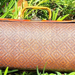 Puede incluir: Un bolso rectangular de mimbre marrón con un patrón de tejido texturizado, un marco de madera lisa, un asa amarilla arqueada y un cierre metálico centrado en el borde superior del bolso, mostrando un diseño estructurado.