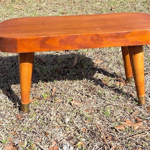 Puede incluir: Un pequeño taburete de madera de estilo moderno de mediados de siglo con acabado marrón rojizo. El taburete tiene una parte superior ovalada y tres patas cónicas con pies de metal color latón. Mide unos 46 cm de largo.