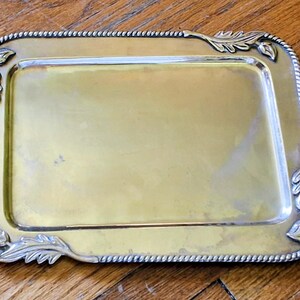 May include: A rectangular brass tray with a smooth, polished centre surface. The edges of the tray feature a decorative rope-like border and raised metallic leaf motifs at each of the four corners, providing a distinct frame for the flat base.