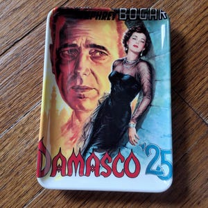 Pode incluir: Bandeja retangular de cerâmica com um design de cartaz de filme vintage. A bandeja apresenta Humphrey Bogart e uma mulher em um vestido preto, com o texto "DAMASCO 25" em vermelho e azul. O fundo é uma mistura de amarelo, laranja e azul.