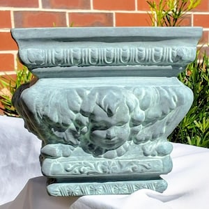 Peut inclure: Un pot de fleurs carré bleu-vert clair orné d'un bas-relief représentant un visage de chérubin entouré de feuillage sur la face avant. Les bords supérieur et inférieur présentent un motif sculpté répétitif et une finition mate.