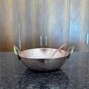 May include: A copper-coloured bowl with a hammered texture and brass handles. A matching spoon rests inside the bowl. The bowl is designed for serving food.