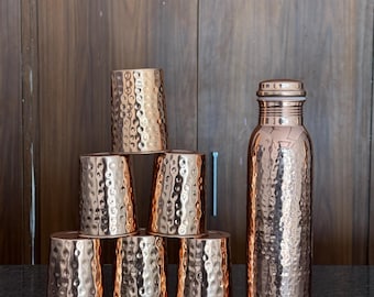 Puede incluir: Una botella de agua de cobre y seis tazas de cobre a juego con una textura martillada. La botella tiene una tapa de rosca. Las tazas están apiladas en forma de pirámide. Los artículos se exhiben sobre una superficie negra.
