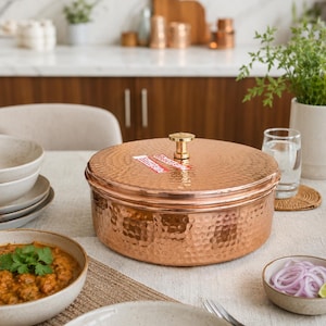 May include: A copper cooking pot with a hammered texture and a matching lid. The pot has a gold-coloured handle on top. The pot is on a table with other dishes and food.
