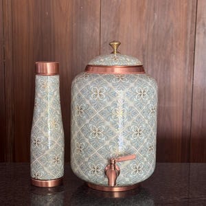 May include: A copper water bottle and a matching copper water dispenser with a spigot. Both items feature a light blue and white floral pattern with gold accents. The dispenser has a removable lid with a small gold knob and a copper base.