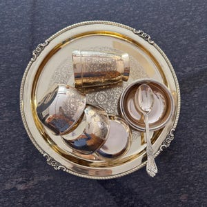 Puede incluir: Un juego de servir dorado con una bandeja redonda con borde ornamentado, una taza peque&ntilde;a, tres cuencos peque&ntilde;os y una cuchara. El juego tiene patrones florales detallados y est&aacute; dispuesto sobre una superficie oscura.