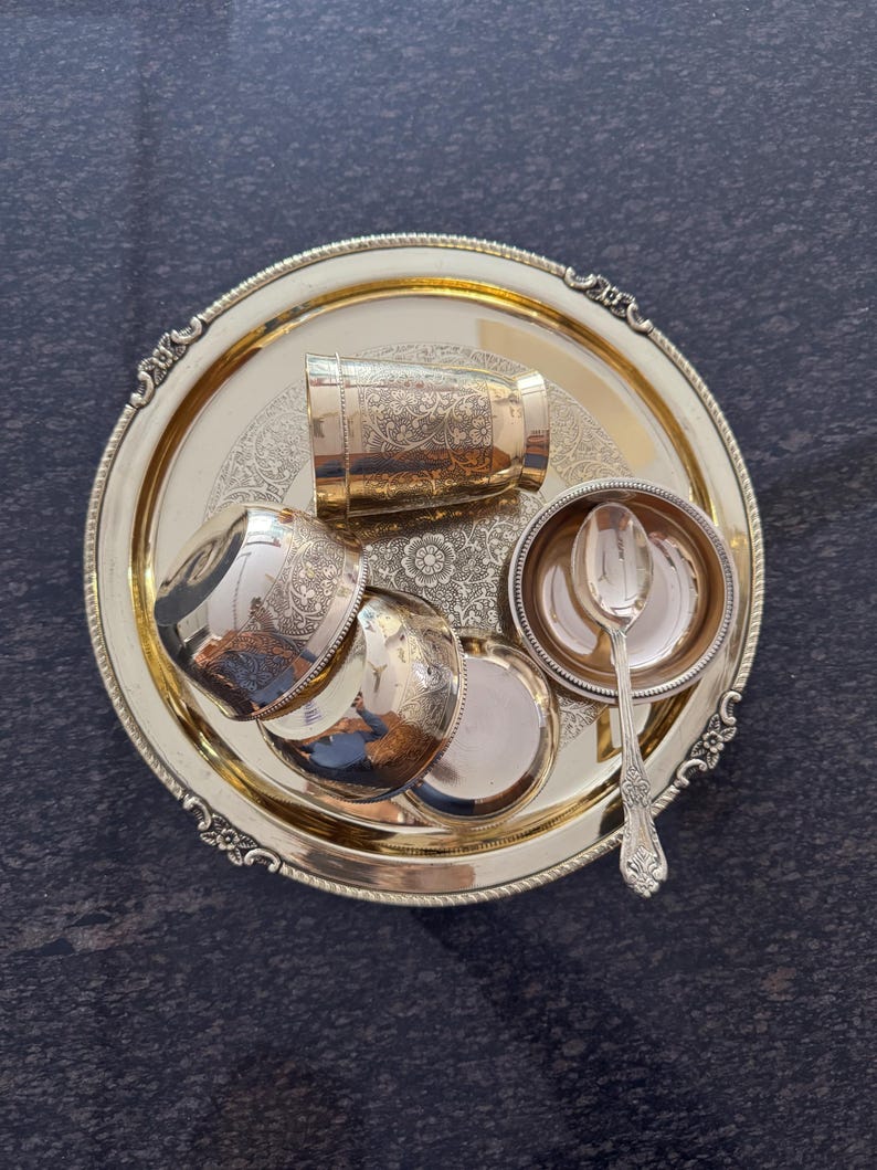 Puede incluir: Un juego de servir dorado con una bandeja redonda con borde ornamentado, una taza peque&ntilde;a, tres cuencos peque&ntilde;os y una cuchara. El juego tiene patrones florales detallados y est&aacute; dispuesto sobre una superficie oscura.