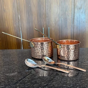 Puede incluir: Dos cacerolas de cobre con mangos largos y dos cucharas de cobre descansan sobre una encimera oscura. Las cacerolas tienen una textura martillada. Las cucharas y los mangos tienen un acabado brillante. El fondo es un armario de madera.