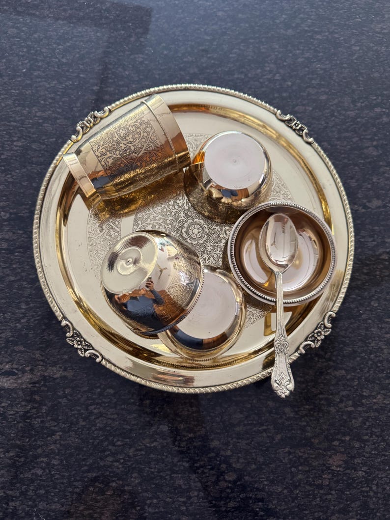Puede incluir: Un juego de servir de metal dorado sobre una bandeja redonda. El juego incluye una taza alta con dise&ntilde;os florales grabados, dos tazas peque&ntilde;as, un cuenco peque&ntilde;o con una cuchara y dos tapas. La bandeja y el juego tienen detalles ornamentados.