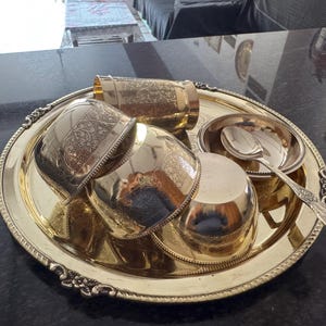 Puede incluir: Una bandeja de servicio de metal dorado con detalles florales ornamentados contiene varias piezas a juego. Estos incluyen una taza alta, cuencos peque&ntilde;os y una cuchara. El conjunto tiene una apariencia antigua y probablemente se utiliza para servir alimentos o bebidas.