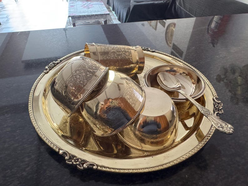 Puede incluir: Una bandeja de servicio de metal dorado con detalles florales ornamentados contiene varias piezas a juego. Estos incluyen una taza alta, cuencos peque&ntilde;os y una cuchara. El conjunto tiene una apariencia antigua y probablemente se utiliza para servir alimentos o bebidas.
