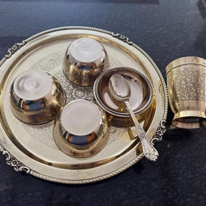 Puede incluir: Un juego de servir dorado con una bandeja decorativa, cuencos peque&ntilde;os y una taza. La bandeja tiene un borde adornado y contiene tres cuencos peque&ntilde;os, un cuenco peque&ntilde;o con una cuchara y una taza con dise&ntilde;os grabados. El juego es probablemente para servir comida o bebidas.