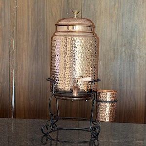 May include: A copper water dispenser with a hammered texture, a tap, and a lid. It sits on a black metal stand with a matching cup. The dispenser is designed for serving beverages.
