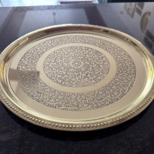 Puede incluir: Una bandeja redonda de metal dorado con un intrincado dise&ntilde;o floral grabado en la superficie. La bandeja tiene un borde con cuentas y un acabado pulido, adecuado para servir o exhibir.
