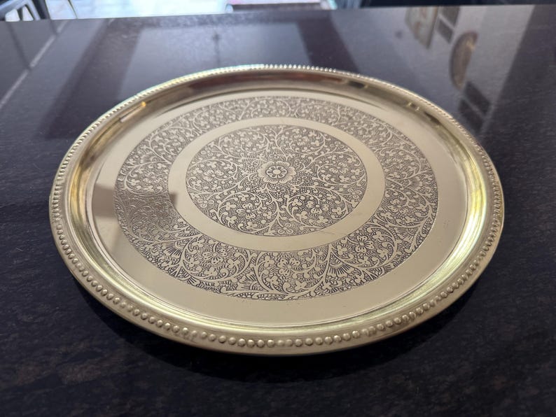 Puede incluir: Una bandeja redonda de metal dorado con un intrincado dise&ntilde;o floral grabado en la superficie. La bandeja tiene un borde con cuentas y un acabado pulido, adecuado para servir o exhibir.