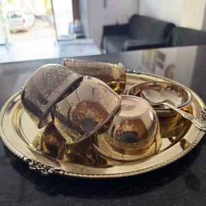 Puede incluir: Una bandeja de metal dorada con un juego de tazas a juego, un cuenco peque&ntilde;o y una cuchara. Las tazas y la bandeja tienen dise&ntilde;os grabados y ornamentados. El conjunto est&aacute; dispuesto sobre una superficie oscura.