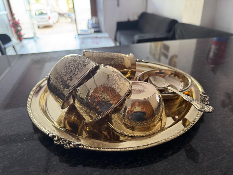 Puede incluir: Una bandeja de metal dorada con un juego de tazas a juego, un cuenco peque&ntilde;o y una cuchara. Las tazas y la bandeja tienen dise&ntilde;os grabados y ornamentados. El conjunto est&aacute; dispuesto sobre una superficie oscura.