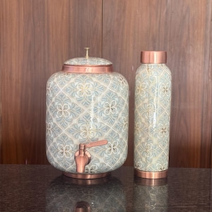 Puede incluir: Un dispensador de agua cilíndrico con tapa y grifo de cobre, junto a una botella de agua alta a juego. Ambos artículos presentan un patrón de azulejos florales en azul claro y blanco con detalles en cobre en los bordes superior e inferior.