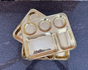 Puede incluir: Una pila de tres platos divididos de metal dorado. Cada plato tiene forma rectangular con múltiples compartimentos de varios tamaños, incluidas secciones redondas y rectangulares. Los platos están apilados sobre una superficie azul oscuro.