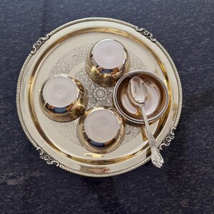 Puede incluir: Una bandeja de metal dorada con cuatro cuencos peque&ntilde;os y una cuchara. La bandeja tiene un dise&ntilde;o floral ornamentado y un borde decorativo. Los cuencos y la cuchara son de color plateado. La bandeja est&aacute; sobre una superficie oscura.