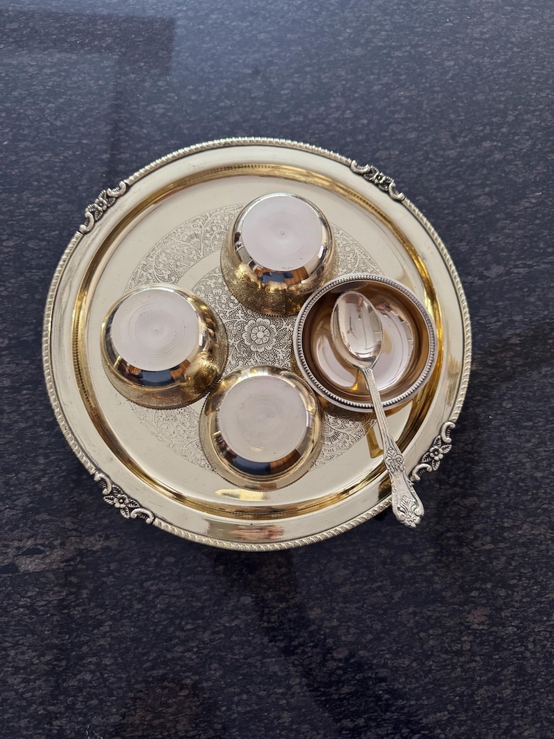 Puede incluir: Una bandeja de metal dorada con cuatro cuencos peque&ntilde;os y una cuchara. La bandeja tiene un dise&ntilde;o floral ornamentado y un borde decorativo. Los cuencos y la cuchara son de color plateado. La bandeja est&aacute; sobre una superficie oscura.