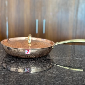 Op de afbeelding: Koperkleurige koekenpan met een deksel en een goudkleurige handgreep. De pan heeft een gehamerde textuur en een goudkleurige handgreep. Het deksel heeft een goudkleurige handgreep. De pan staat op een donkere ondergrond.