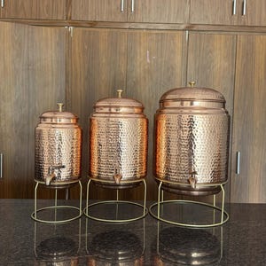 Puede incluir: Tres dispensadores de agua de color cobre con texturas martilladas y boquillas de latón. Cada dispensador tiene una tapa y se asienta sobre un soporte de metal dorado. Los dispensadores varían en tamaño, el más grande a la derecha.
