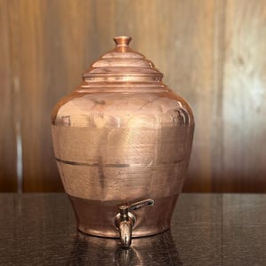 May include: A copper-colored water dispenser with a spigot. The dispenser has a rounded body with a lid and a horizontal band. The spigot is made of a shiny metal. The dispenser is sitting on a dark surface.