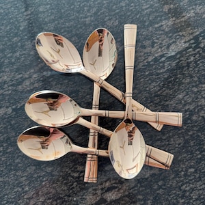 May include: Six copper-colored spoons arranged in a starburst pattern. The spoons have oval bowls and rectangular handles with etched lines. The spoons reflect the surrounding environment, creating a mirrored effect.