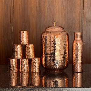 May include: A collection of copper drinkware, including a large hammered water dispenser with a spigot, a water bottle, and six matching cups. The copper items have a textured surface and are arranged on a reflective surface.