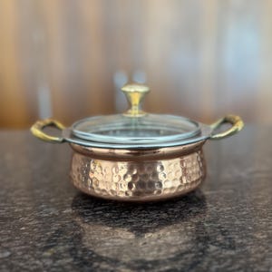 Puede incluir: Una olla de cocina de color cobre con una textura martillada, una tapa de cristal transparente y asas doradas. La olla está diseñada para cocinar y servir alimentos. La olla está sobre una superficie oscura.