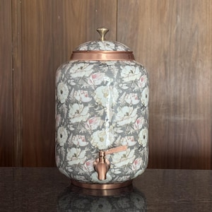 May include: A cylindrical beverage dispenser featuring a grey background with a white and pink floral pattern. The container includes a copper-coloured metal lid with a small knob, a matching copper-coloured spigot, and a copper base rim.