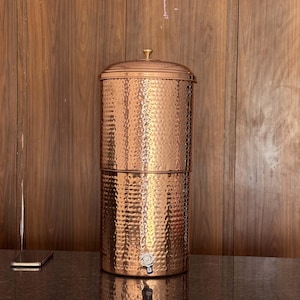 Puede incluir: Filtro de agua de cobre martillado con tapa y grifo. El recipiente cilíndrico tiene una superficie texturizada y un acabado brillante. Se encuentra sobre una superficie oscura, con un fondo de madera.