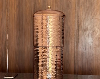 Può includere: Un filtro per l'acqua in rame con una texture martellata. Il contenitore cilindrico ha un coperchio con un piccolo pomello e un rubinetto vicino al fondo. La finitura in rame gli conferisce un aspetto rustico ed elegante.