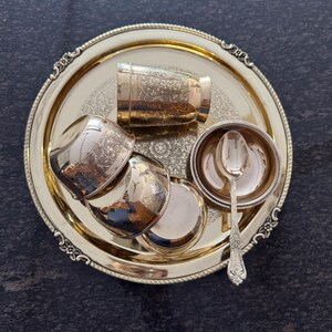 Puede incluir: Un juego de servir de metal dorado, que incluye una bandeja redonda, tazas peque&ntilde;as, un cuenco y una cuchara. La bandeja y las tazas tienen dise&ntilde;os florales ornamentados. El juego est&aacute; dispuesto sobre una superficie oscura y texturizada.