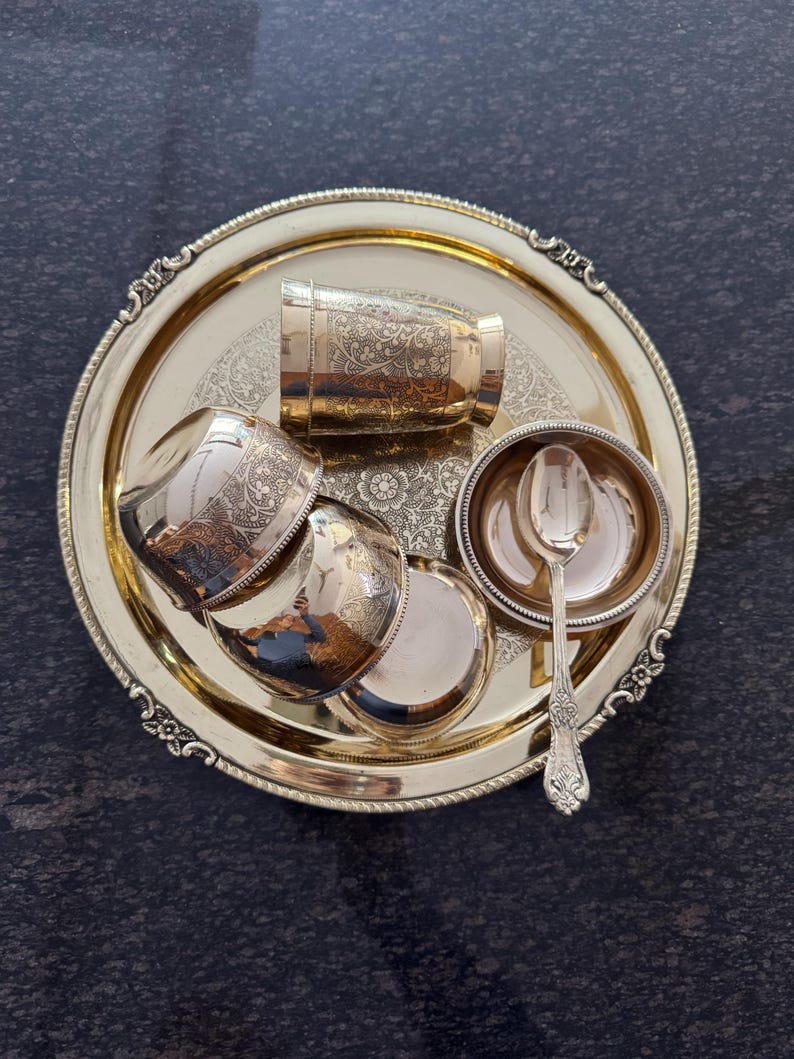 Puede incluir: Un juego de servir de metal dorado, que incluye una bandeja redonda, tazas peque&ntilde;as, un cuenco y una cuchara. La bandeja y las tazas tienen dise&ntilde;os florales ornamentados. El juego est&aacute; dispuesto sobre una superficie oscura y texturizada.