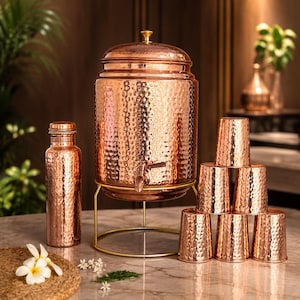 Puede incluir: Un dispensador de agua de cobre martillado con tapa y grifo, sobre un soporte de metal dorado. El juego incluye una botella de cobre martillado a juego y seis vasos de cobre martillado apilados en pirámide sobre una superficie de mármol.